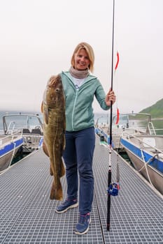 Happy young fisherwoman holding big arctic cod. Norway happy fishing. Happy woman with cod fish in hands, night polar day. Guided fishing concept. Fishing tourism. Active vacation, open sea, ocean fishing.