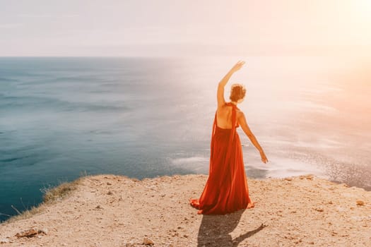 Side view a Young beautiful sensual woman in a red long dress posing on a rock high above the sea during sunrise. Girl on the nature on blue sky background. Fashion photo.