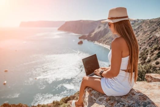 Successful business woman in yellow hat working on laptop by the sea. Pretty lady typing on computer at summer day outdoors. Freelance, travel and holidays concept.