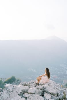 Young woman with a hat in her hand sits on a rocky cliff and looks at the mountains. Back view. High quality photo