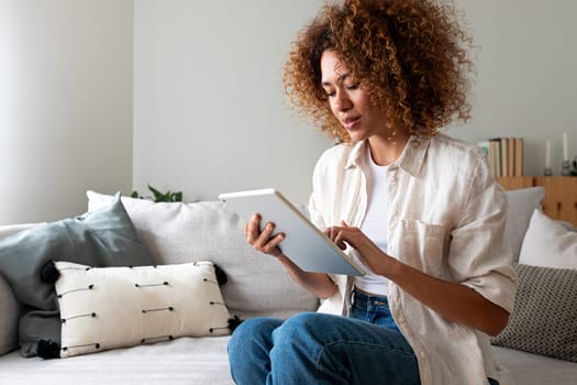 Young multiracial Hispanic woman using digital tablet sitting on the sofa at home living room. Copy space. Lifestyle and technology concept.