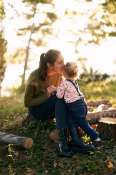 Mom sits on a stump and kisses on the cheek a little girl standing next to her in the forest. High quality photo