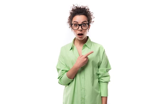 young surprised brunette businesswoman with curly hair dressed in a green shirt points her hand to the side.