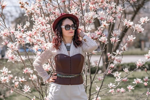 Magnolia park woman. Stylish woman in a hat stands near the magnolia bush in the park. Dressed in white corset pants and posing for the camera