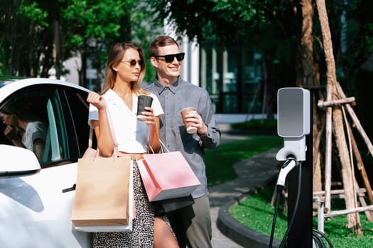 Young couple travel with EV electric car charging in green sustainable city outdoor garden in summer shows urban sustainability lifestyle by green clean rechargeable energy of electric vehicle innards