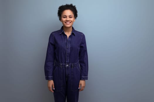 young positive brunette latin woman with afro hair gathered in a ponytail dressed in a blue stylish suit consisting of one-piece jeans and a shirt.