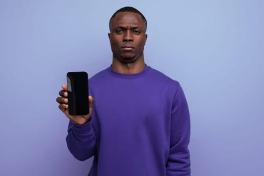authentic handsome young african man in blue sweater holding smartphone vertically with mockup for advertisement on blue background with copy space.