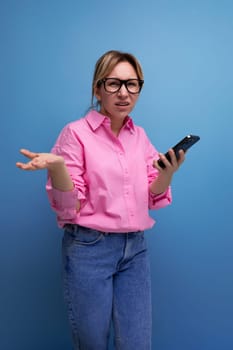 young european office worker woman dressed in a pink shirt confusedly holding a smartphone and shrugging her shoulders.