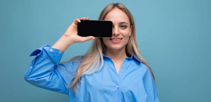 attractive confident blond girl holding a smartphone with a screen at the camera with a blank mockup for a website on a blue background.