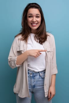 funny european young brunette woman in shirt and jeans pointing finger to the side on blue background.