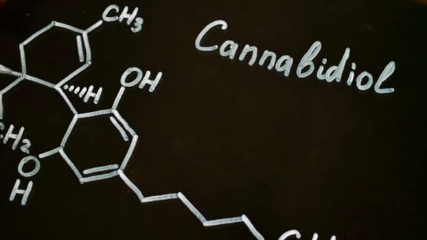 Close-up drawing of the chemical formula of cannabidiol on a black background. A scientist chemist writes in white