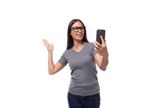 young positive brunette woman with glasses dressed in a gray t-shirt speaks by smartphone video link on a white background with copy space.