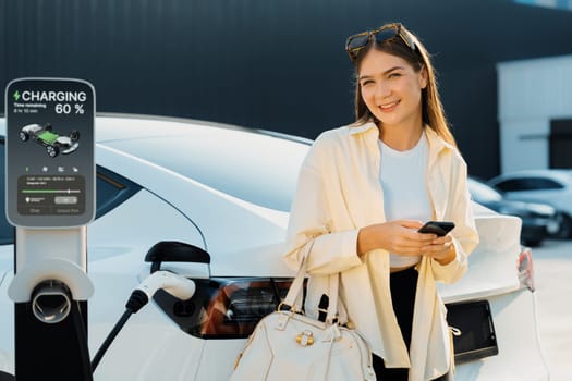 Young woman holding shopping bag and use smartphone to pay for electricity for recharging EV car battery from charging station at city mall parking lot. Modern woman go shopping by eco car. Expedient