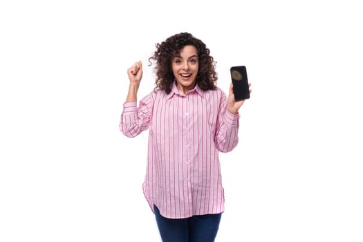 young curly caucasian woman dressed in a striped pink shirt rejoices holding a phone with a mockup.