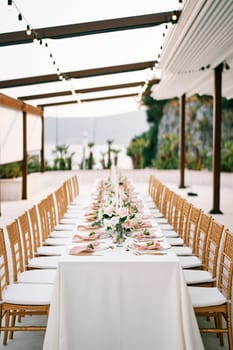 Long festive table with bouquets of flowers under a canopy in the garden, surrounded by soft wicker chairs. High quality photo