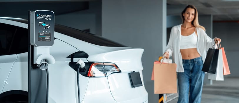 Young woman travel with EV electric car to shopping center parking lot charging in downtown city showing urban sustainability lifestyle by green clean rechargeable energy of electric vehicle innards