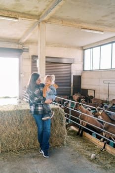 Mother with a little girl in her arms stands leaning on a haystack near a goat paddock. High quality photo