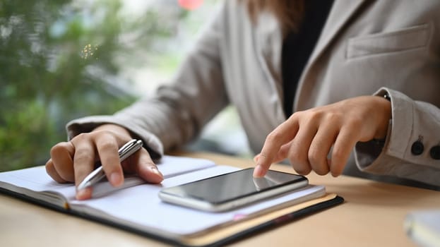 Young businesswoman using mobile phone at office desk and making notes.