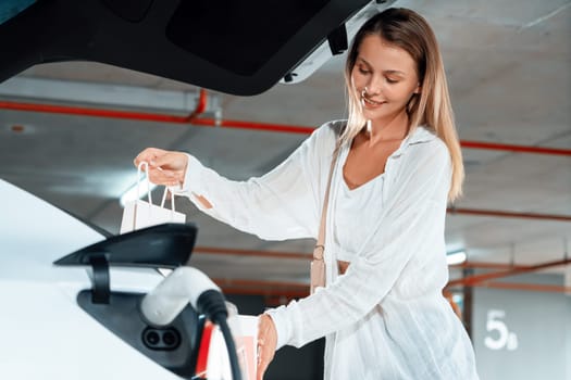 Young woman travel with EV electric car to shopping center parking lot charging in downtown city showing urban sustainability lifestyle by green clean rechargeable energy of electric vehicle innards