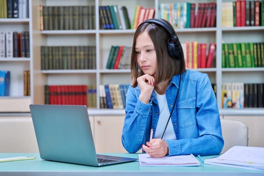 Young female college student in headphones sitting at desk with laptop having online video conference chat, looking at screen talking listening watching learning class, webinar, lesson inside library