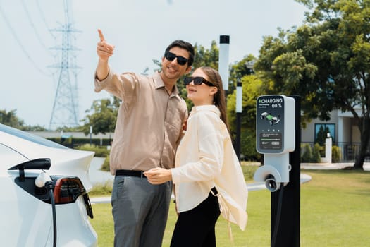 Young couple with coffee pay for electricity while recharge EV car battery at charging station connected to electrical power grid tower as electric industrial for eco friendly car travel. Expedient