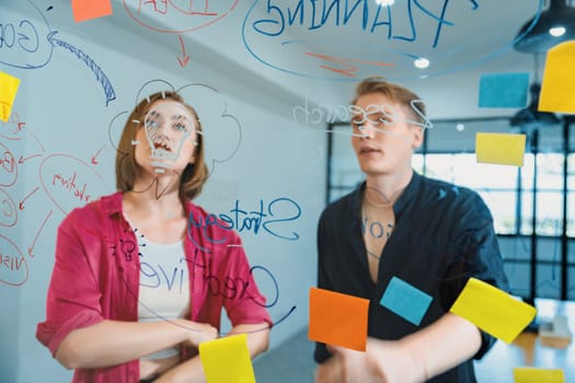 Couple of businesspeople discussing and brainstorming about solving marketing problem in front of glass board at business meeting by using mind map and sticky notes. Working together. Immaculate.