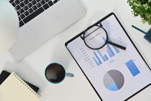 Top view laptop, magnifying glass and financial reports on white table.