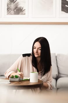 A brunette girl sitting at a desk in the office, making notes in a notebook on work with a mug of tea