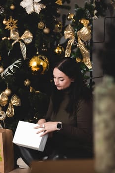 A brunette girl is sitting next to a Christmas tree and looking at a gift