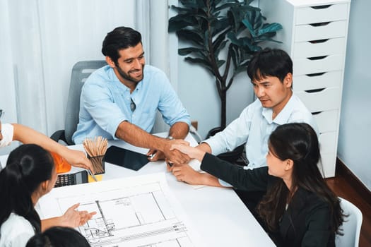 Diverse group of civil engineer and client shakes hand after make successful agreement on architectural project, reviewing construction plan and building blueprint at meeting table. Prudent