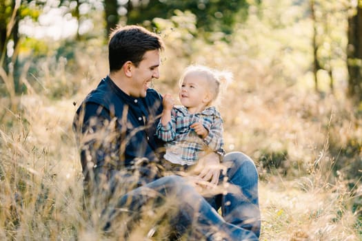 Little girl shows her dad a blade of grass while sitting on his lap on the lawn. High quality photo