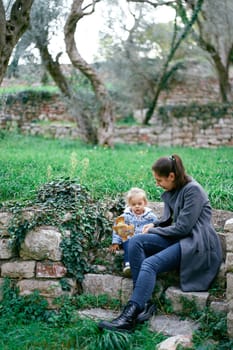 Mom with a little girl sits on stone steps in the garden and shows her a yellow branch. High quality photo