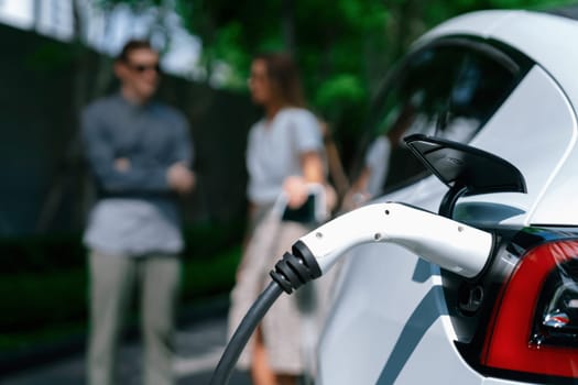 Young couple travel with EV electric car charging in green sustainable city outdoor garden in summer shows urban sustainability lifestyle by green clean rechargeable energy of electric vehicle innards