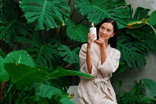 Tropical and exotic spa garden with bathtub in modern hotel or resort with woman in bathrobe holding beauty skincare product while enjoying leisure lush with greenery foliage background. Blithe