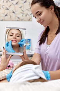 Beautiful woman looks at herself in the mirror after procedure lip augmentation in beauty medical clinic.