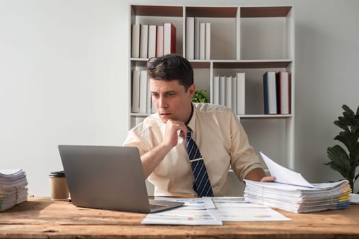 Unhappy young businessman feeling bored and stressed at work looking at laptop with hopeless expression while sitting in office.