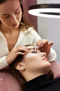 beautician with tweezers makes eyelash extension for young woman in beauty salon.