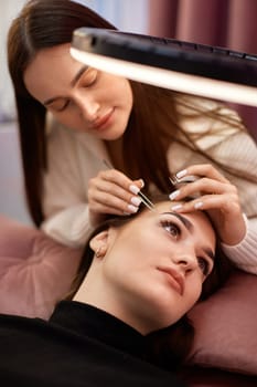 professional beautician with tweezers makes eyelash extension for beautiful woman under ring light lamp, top view
