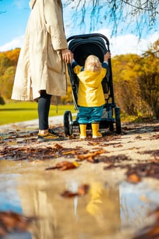 Sun always shines after the rain. Small blond infant boy wearing yellow rubber boots and yellow waterproof raincoat walking in puddles, pushing stroller in city park, holding mother's hand after rain