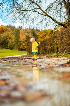 Sun always shines after the rain. Small bond infant boy wearing yellow rubber boots and yellow waterproof raincoat walking in puddles in city park on sunny rainy day