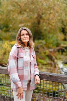 Portrait of cute young woman in casual wear in autumn, standing on bridge against background of an autumn Park and river. Pretty female walking in Park in golden fall. Copy space. smiling girl in the park standing on wooden bridge and looking at the camera in autumn season