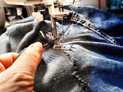 Sewing machine and blue jeans fabric. A woman's hand and fingers next to a sewing machine needle when sewing up tear in denim