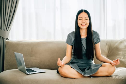 Asian woman meditates in lotus pose on sofa with laptop finding balance. Businesswoman embraces relaxation and mindfulness while studying online. Portrait of a smiling harmonious student.