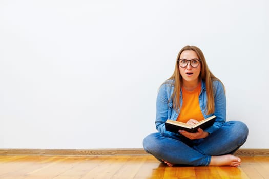 A surprised, positive young woman with glasses sits on the floor against a white wall holding a book