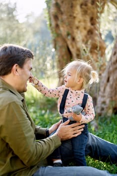 Little girl sits on her father lap with a yellow branch in her hand and tickles his nose. High quality photo