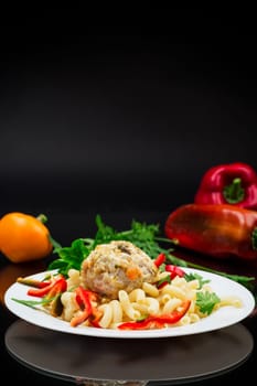 cooked pasta with meatballs, peppers and herbs with spices in a plate, isolated on a black background