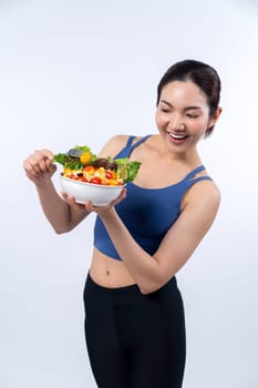 Young sporty Asian woman in sportswear holding salad bowl fill with vibrant of fruit and vegetable. Natural youthful and fit body lifestyle with balance nutrition on isolated background. Vigorous