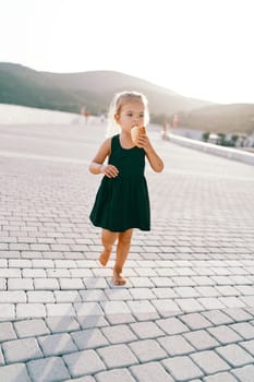 Little girl eats a bun while walking barefoot on a tiled road. High quality photo