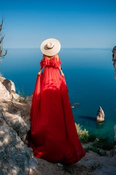 Red dress sea woman. Happy woman with flowing hair in a long flowing red dress stands on a rock near the sea. Travel concept, photo session at sea