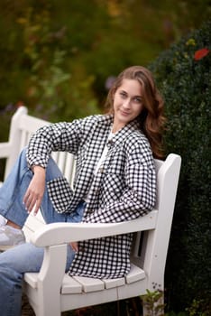 Beautiful teen blonde girl sits on a bench in an autumn park . attractive young smiling girl sitting on a bench in an autumn park. Fall Fashion Serenity: Casual Elegance in the Park